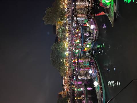       Dozens of tourists ride small wooden boats decorated with colorful lanterns on a river under the night sky, city lights and trees lining the banks.
  