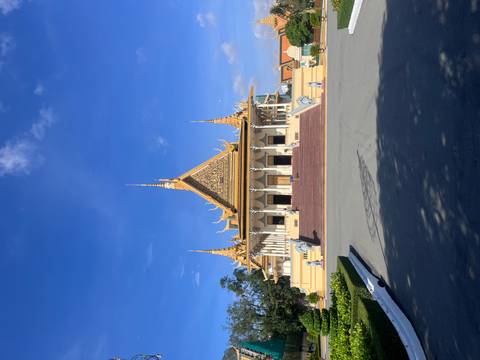       Grand golden-roofed Khmer palace building with ornate spires sits under a clear blue sky, framed by a wide stone courtyard.
  
