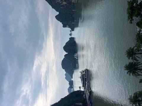       Karst islands rise from calm green waters of a wide bay where cruise boats are anchored under a cloudy sky.
  