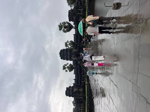       Visitors with umbrellas walk along a wet stone causeway toward the towering sandstone spires of Angkor Wat on a rainy day.
  