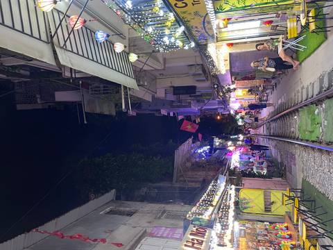       Nightview of Hanoi Train Street with railway tracks flanked by lit cafés, lanterns and people enjoying drinks.
  