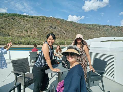       Small group of travellers enjoy sunshine on the upper deck of a Rhine river cruise boat with hillsides behind.
  