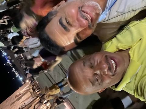       Close-up selfie of two men smiling at a crowded night event with blurred people and lights in the background.
  