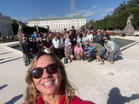       Large tour group posing for a selfie in front of the ornate Mirabell Palace gardens in Salzburg on a sunny day.
  