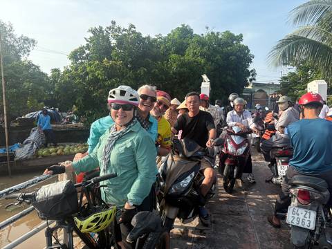       Cyclists wearing helmets queue on a small ferry with motorbikes in tropical sunlight.
  