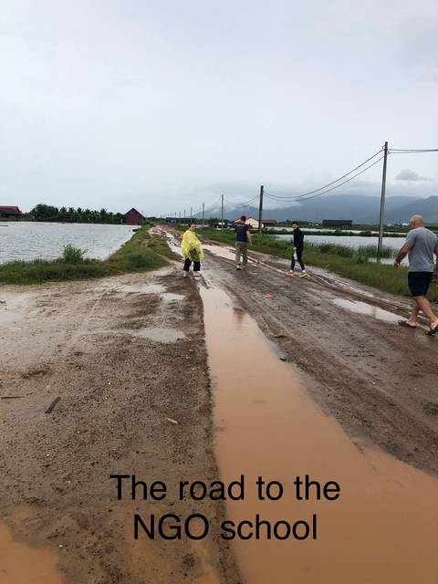       Travelers walk along a muddy rural road lined with rice paddies under grey skies.
  