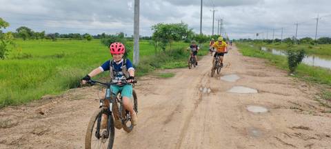       Children and adults ride mountain bikes down a rural dirt track flanked by green rice fields.
  