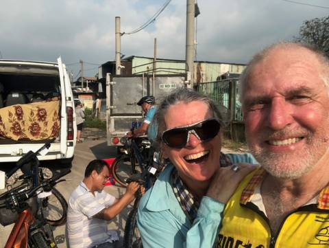       Close-up selfie of a laughing couple with support vehicle and bikes in the background.
  