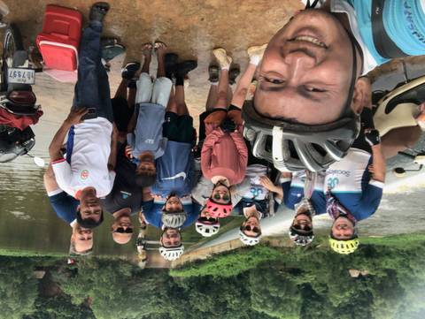       Large group of cyclists pose together on a riverbank with scooters and locals nearby.
  