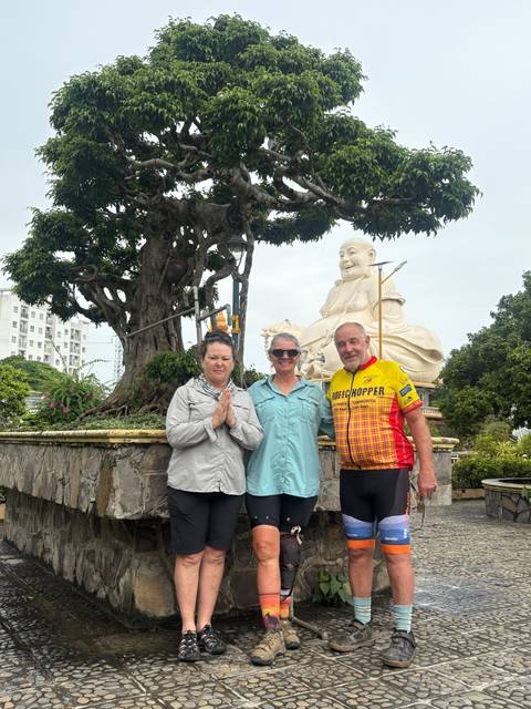       Three travelers pose before a giant smiling Buddha statue and sacred tree.
  