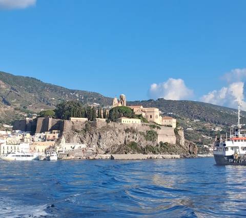       Fortified hilltop town of Lipari rising above rocky cliffs and blue Tyrrhenian Sea as boats approach.
  