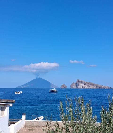       Distant view of steaming Stromboli volcano and rugged offshore islet against clear blue sky.
  