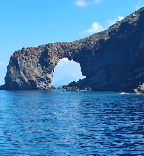       Natural rock arch rising from blue Mediterranean waters with distant volcanic peak framed in opening.
  