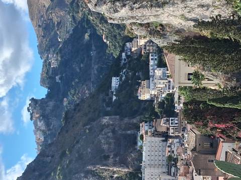       Steep green hills with clustered hillside houses and rocky cliffs around Taormina on a sunny day.
  