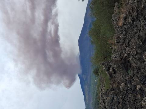       Dark plume of ash rising from snow-capped Mount Etna above rugged lava fields and scrubland.
  