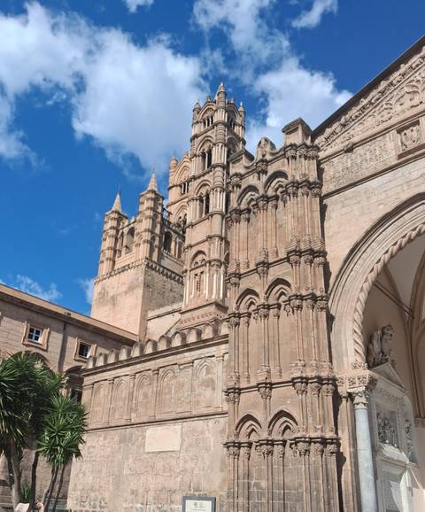       Detailed facade of Palermo Cathedral with its ornate towers and arches under bright blue sky.
  