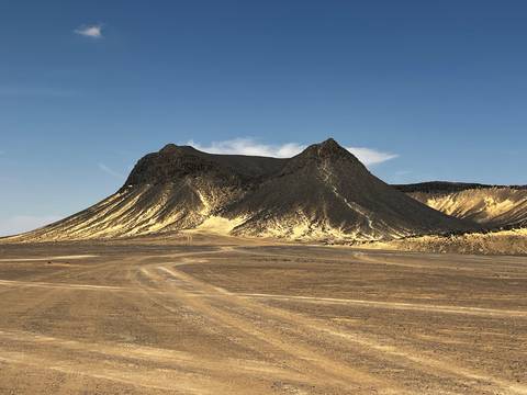       Dark volcanic cone with light sand streaks stands against a clear blue sky in Egypt’s Black Desert.
  