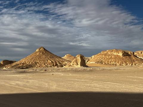       Series of ochre desert hills and peaks under mixed clouds with flat sandy foreground.
  