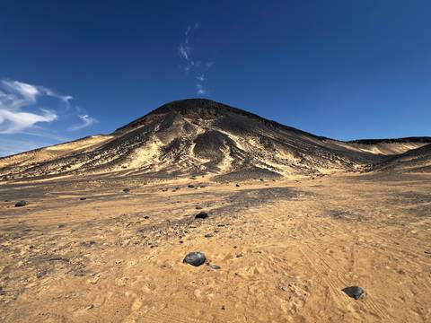       Symmetrical black volcanic hill rises above sandy plain dotted with dark boulders under clear sky.
  