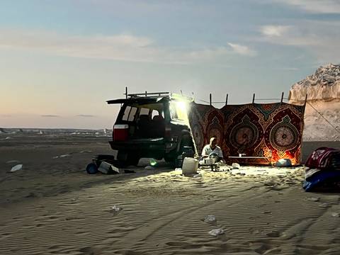       Bedouin guide prepares tea beside 4x4 and patterned carpet under lantern light in remote desert night camp.
  