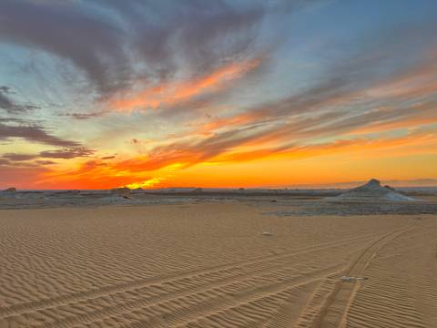       Fiery sunset paints streaks of orange and pink across the sky above rippled desert sand.
  