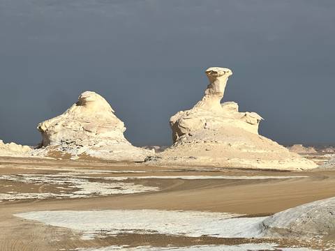       Twin chalk towers rise from a sandy desert plain under a clear blue sky.
  