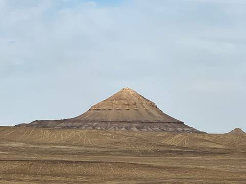       Isolated conical hill rises from flat desert terrain against pale morning sky.
  