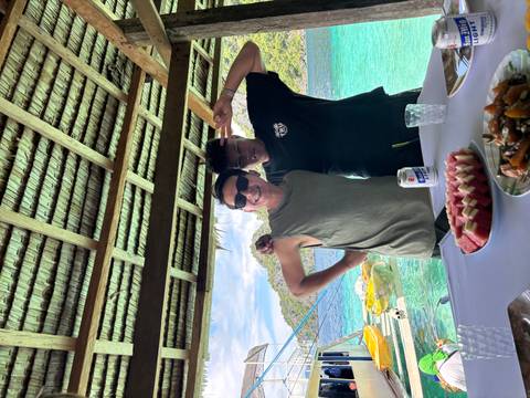       Two friends pose inside a thatched hut overlooking turquoise sea with tropical islands in the background.
  