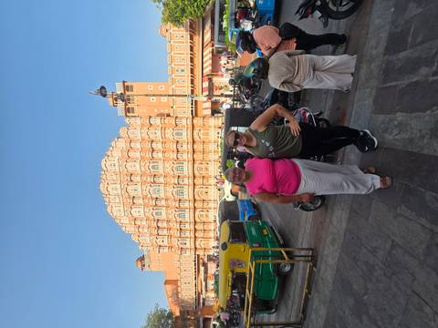       Travellers pose on busy street with the ornate façade of Jaipur’s Hawa Mahal rising behind
  