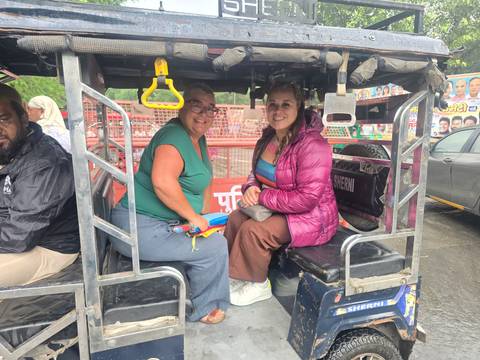       Travellers sit in the back of an open e-rickshaw on a rainy street, wearing casual jackets
  