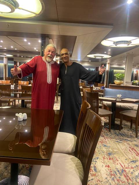       Two friends in traditional robes smile and embrace inside a ship restaurant with polished wood tables.
  