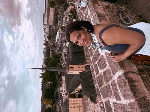       Woman smiling on stone battlement overlooking old European town with church spire and green hills in background.
  