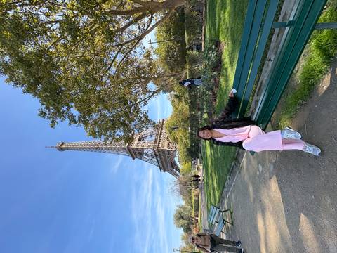       Visitor in pink outfit sitting on a park bench with the Eiffel Tower rising behind against a blue sky.
  