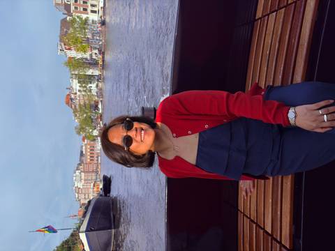       Woman cruising on a city canal boat with historic waterfront buildings in soft light behind her.
  