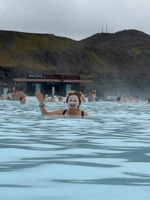       Visitors bathing in milky blue geothermal waters of the Blue Lagoon, one woman waving with silica mask on face.
  