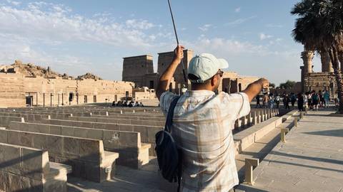       A tour guide gestures toward the ancient stone temples of Philae as rows of empty seats await visitors.
  