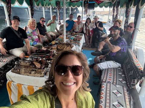       A smiling tour group poses on a cushioned boat deck displaying colorful local handicrafts on the Nile.
  
