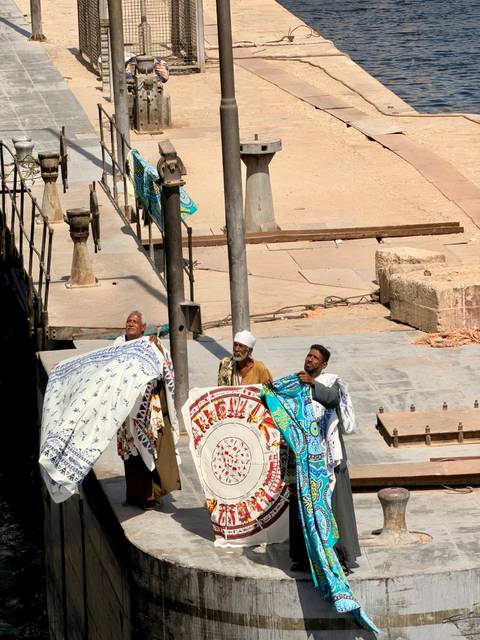       Three vendors holding colourful patterned blankets on a riverside pier under strong sun
  