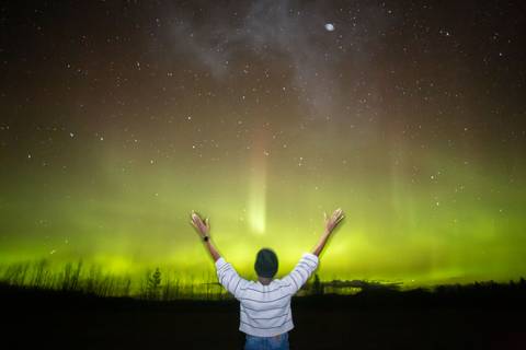       Back view of traveler raising hands in awe at a shimmering green aurora overhead
  