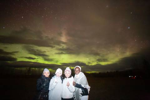       Small group bundled in winter gear smiling with faint aurora and cloud-filled night sky behind
  