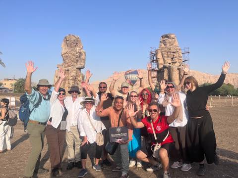       Tour group cheering and waving in front of the Colossi of Memnon with a hot-air balloon behind
  