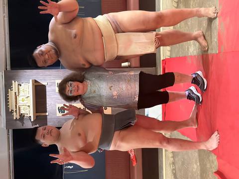      Visitor posing with two sumo wrestlers on a red mat inside a training arena.
  