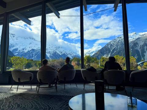       Travelers relax in lounge chairs gazing through large windows at snow-covered mountains and dramatic skies
  