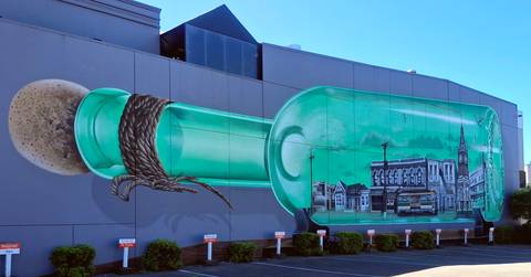       Large street-art mural of a green bottle containing a town scene painted on the side of a modern building
  