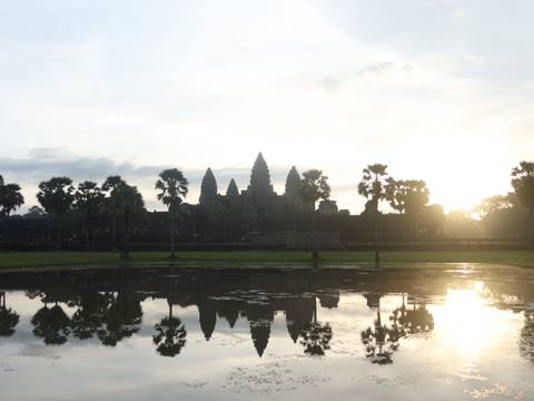       Silhouetted spires of Angkor Wat emerge against a hazy sunrise, reflected in a lotus pond.
  