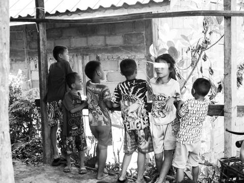       A black-and-white image of village children peer through a wooden railing, curious and contemplative.
  