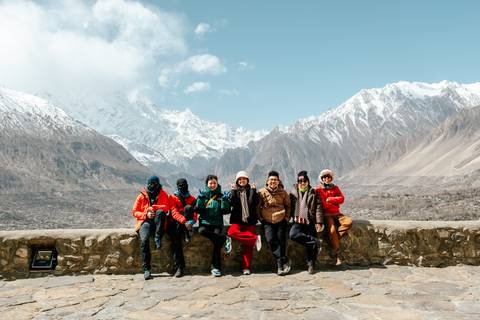       A joyful group of travellers sits on a stone wall with snow-capped peaks of Hunza Valley behind them.
  