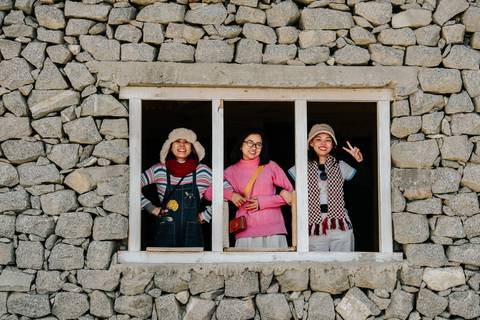       Three women smile through a stone-framed window of a traditional mountain house.
  