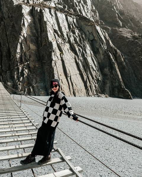       A woman poses on the narrow wooden planks of the Hussaini Suspension Bridge spanning a rocky gorge.
  