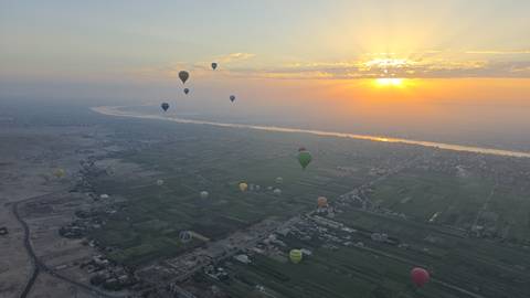       Dozens of colorful hot-air balloons drift above the Nile valley at sunrise casting light over fields and river.
  